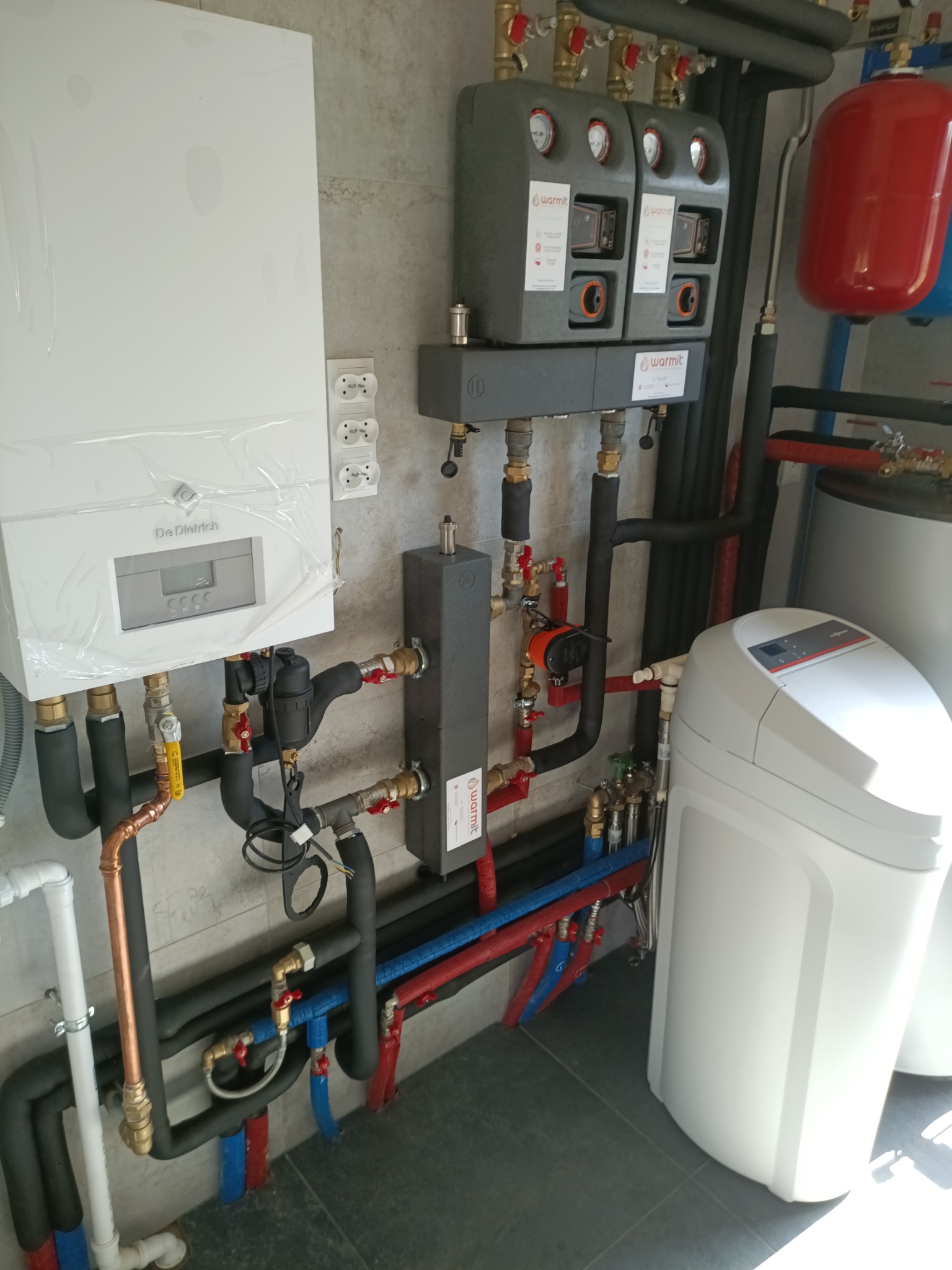 Kotłownia z piecem De Dietrich, zbiornikiem wyrównawczym, zmiękczaczem wody i licznymi rurami miedzianymi oraz z tworzyw sztucznych.