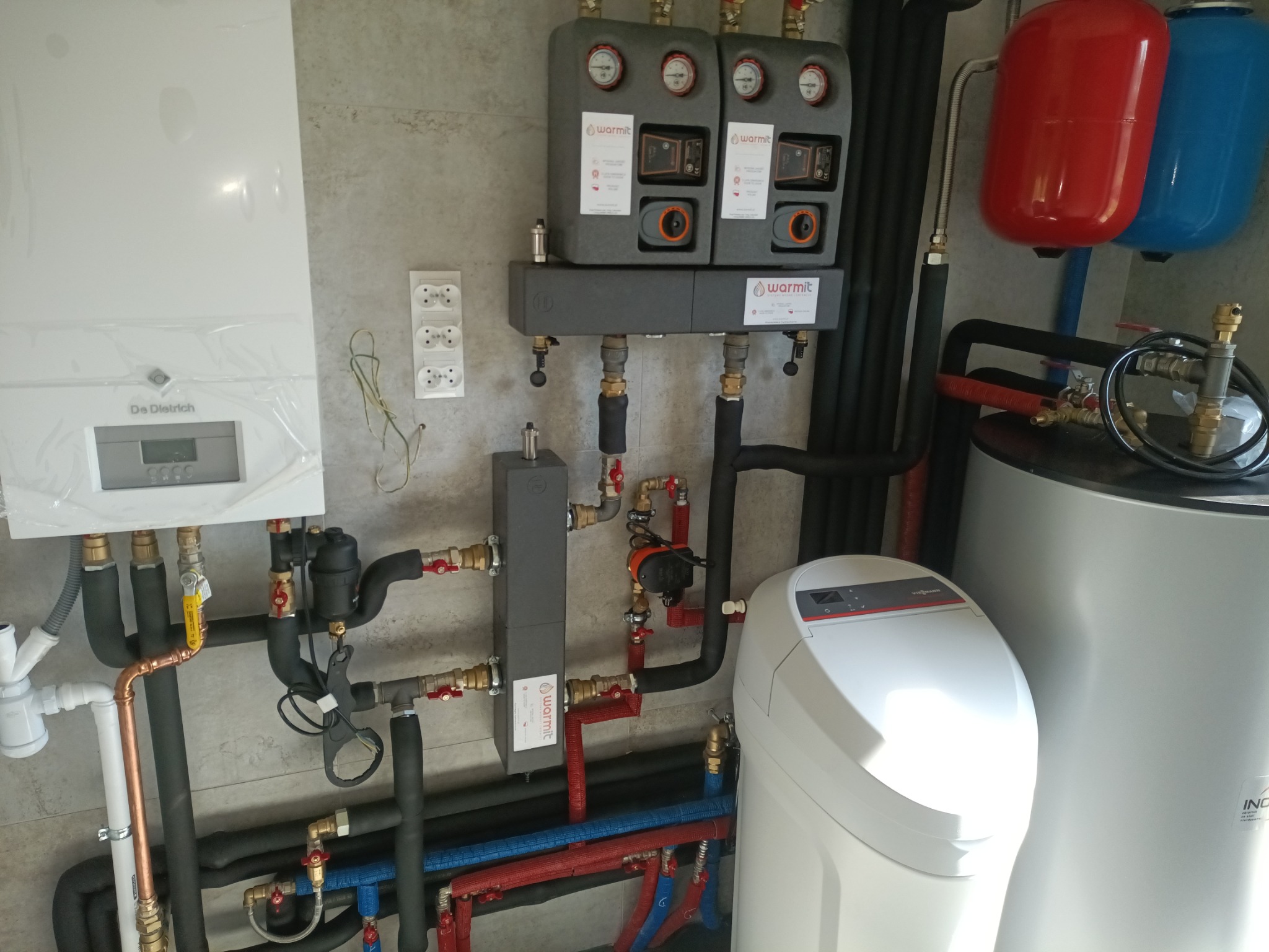 Zestaw instalacji grzewczej: piec De Dietrich, rozdzielacze Warmit, czarne rury izolowane, zbiornik buforowy Viessmann i zmiękczacz wody, na tle ściany z szarą płytką.
