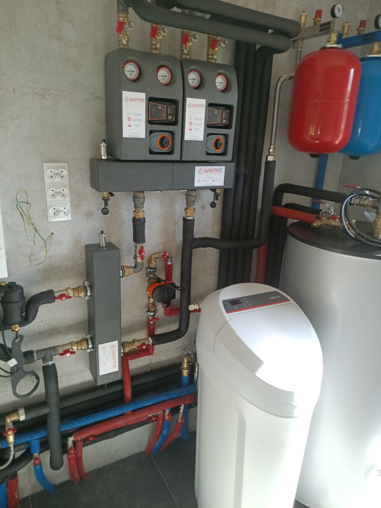 Złożona instalacja grzewcza z kotłem, zasobnikiem i filtrem wody, rurami w izolacji, manometrami i zaworami, zamontowana na ścianie wyłożonej płytkami.