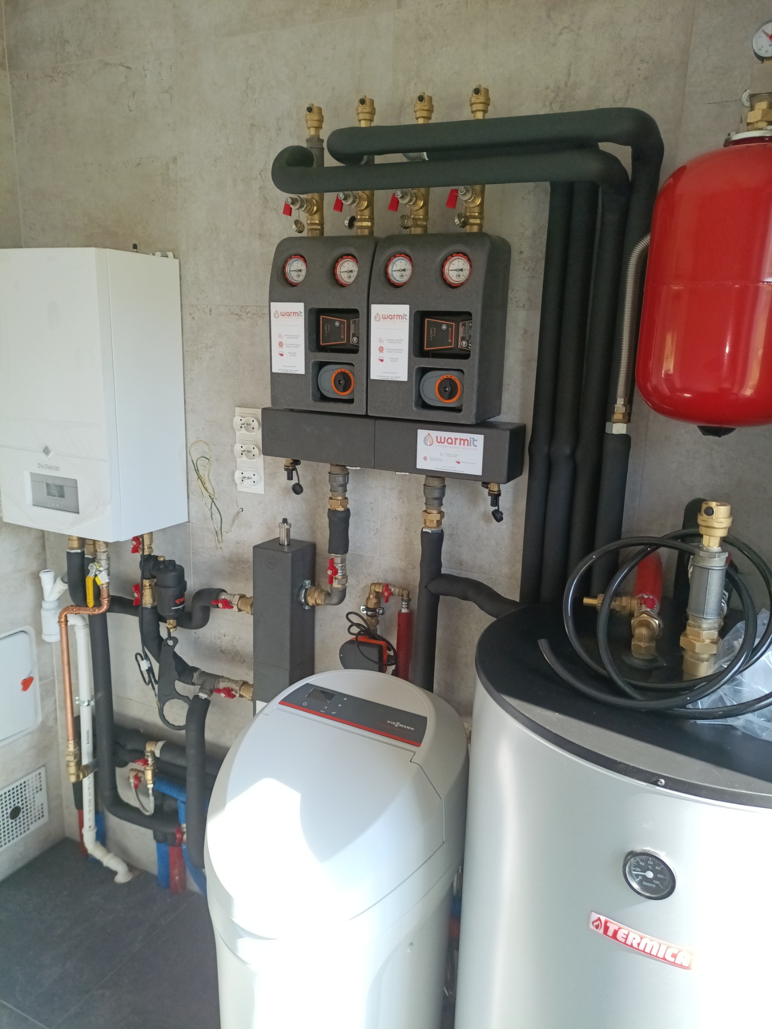 Kompletna kotłownia z piecem gazowym, zasobnikiem, grupą pompową Warmit i filtrem. Widoczne rury, zawory i manometry. Nowoczesna instalacja grzewcza w pomieszczeniu.