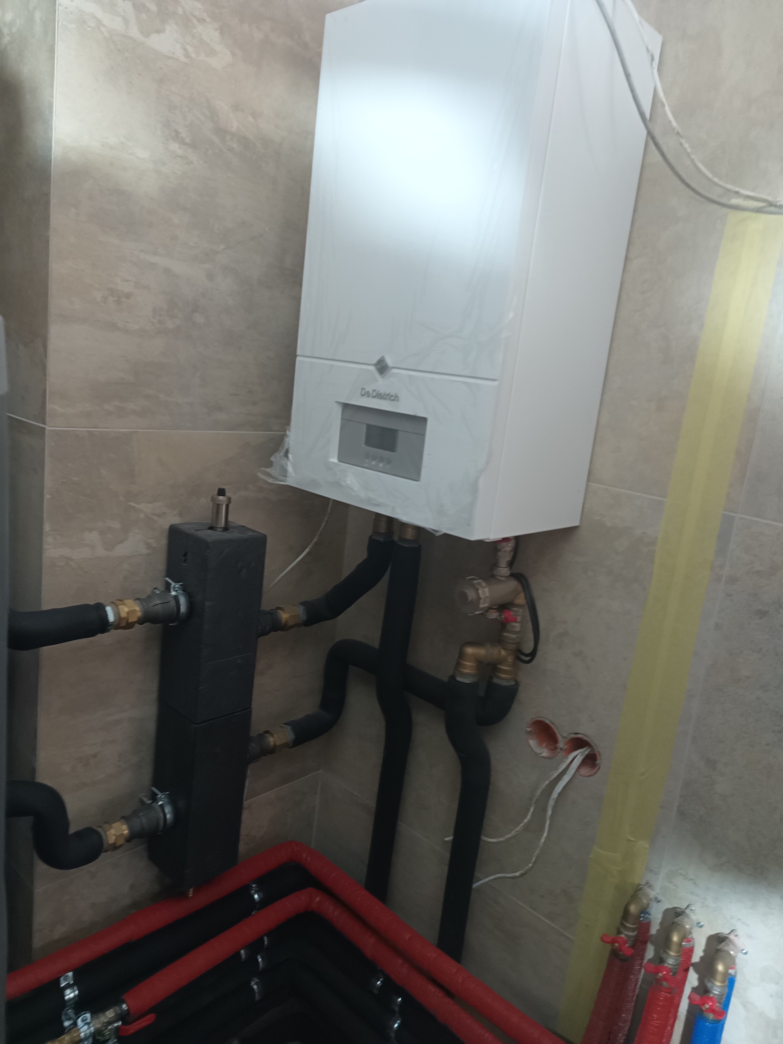 Biały piec gazowy De Dietrich zamontowany na beżowej ścianie z widocznymi czarnymi rurami, elementami instalacji i fragmentem czerwono-czarnego ogrzewania podłogowego.