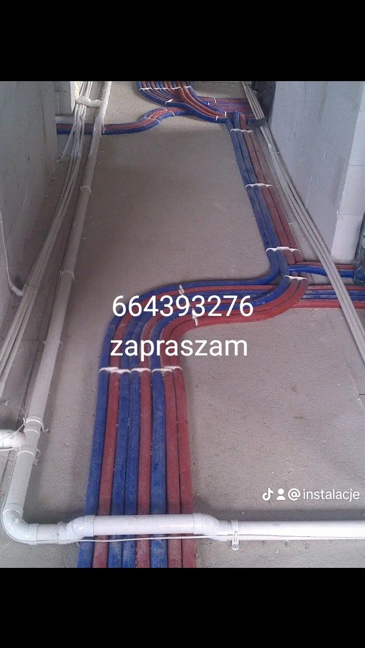 Instalacja ogrzewania podłogowego z rur PEX w kolorze czerwonym i niebieskim, ułożonych równolegle na szarej posadzce w surowym stanie. Widoczne białe rury instalacji wodnej.