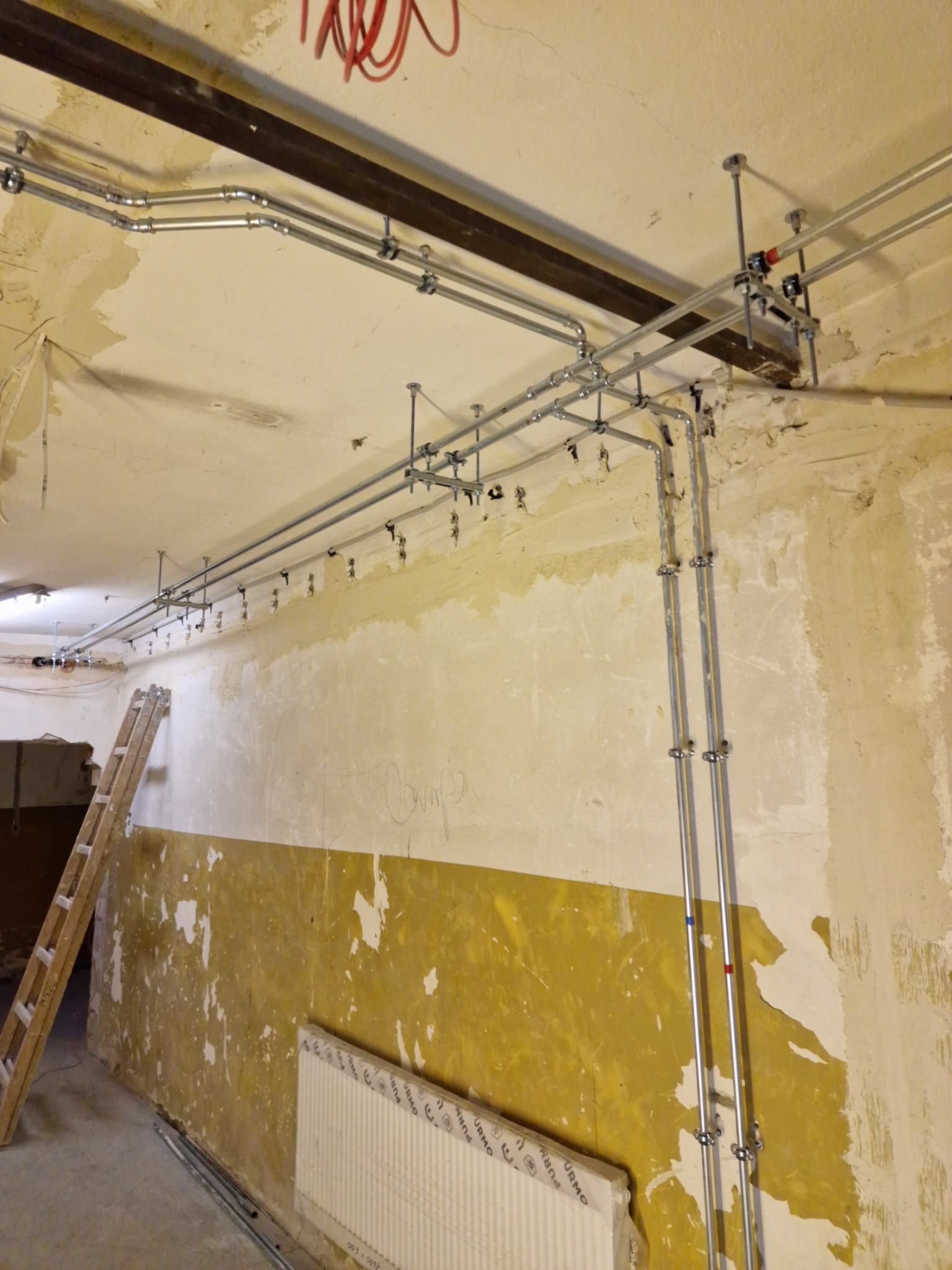 Metalowe rury instalacji podwieszone do sufitu w pomieszczeniu w trakcie remontu, widoczna drabina oparta o ścianę z resztkami farby i fragment grzejnika w folii.