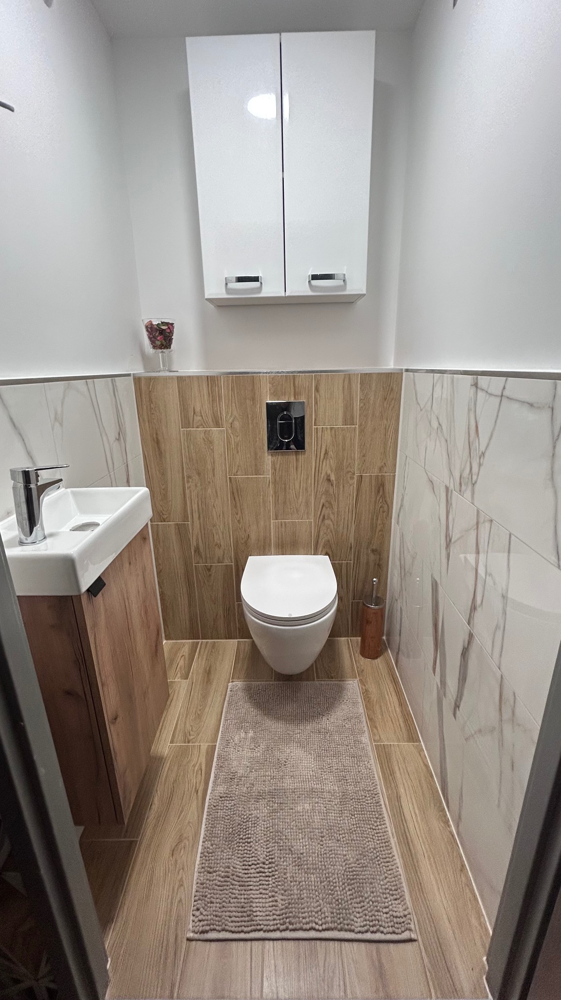 Nowoczesna toaleta: drewnopodobne i marmurowe płytki, podwieszana miska WC, szafka z umywalką. Minimalistyczny design, beżowy dywanik. Górna szafka z białymi frontami.