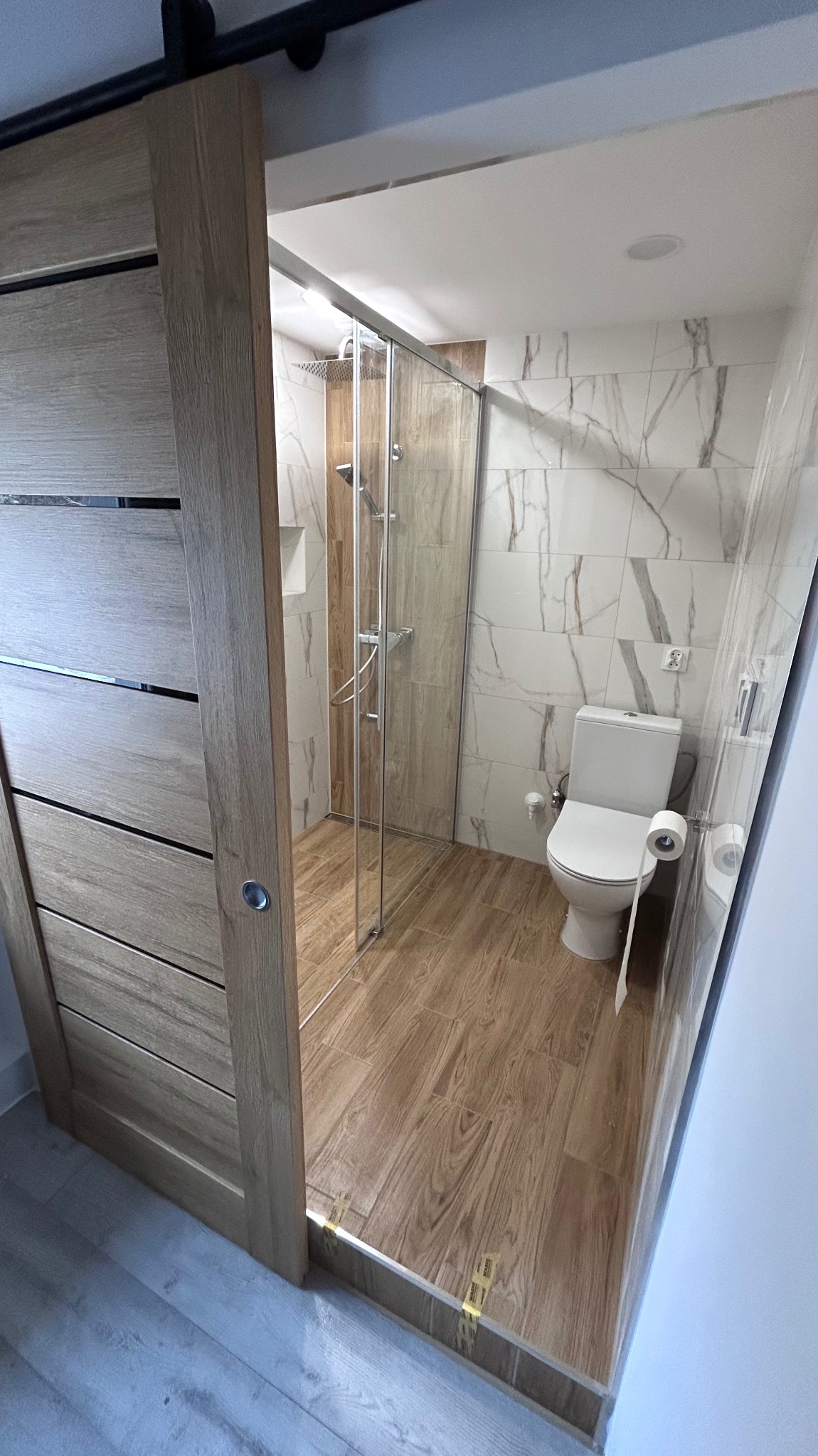 Nowoczesna łazienka po remoncie: kabina prysznicowa ze szklanymi drzwiami, toaleta i marmurowe płytki. Drewnopodobna podłoga i przesuwne drzwi z drewnianym wykończeniem.