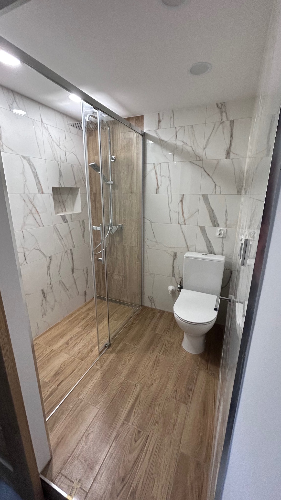 Nowoczesna łazienka z kabiną prysznicową typu walk-in, wykończona płytkami imitującymi marmur i drewno. Widoczna toaleta i minimalistyczne oświetlenie.