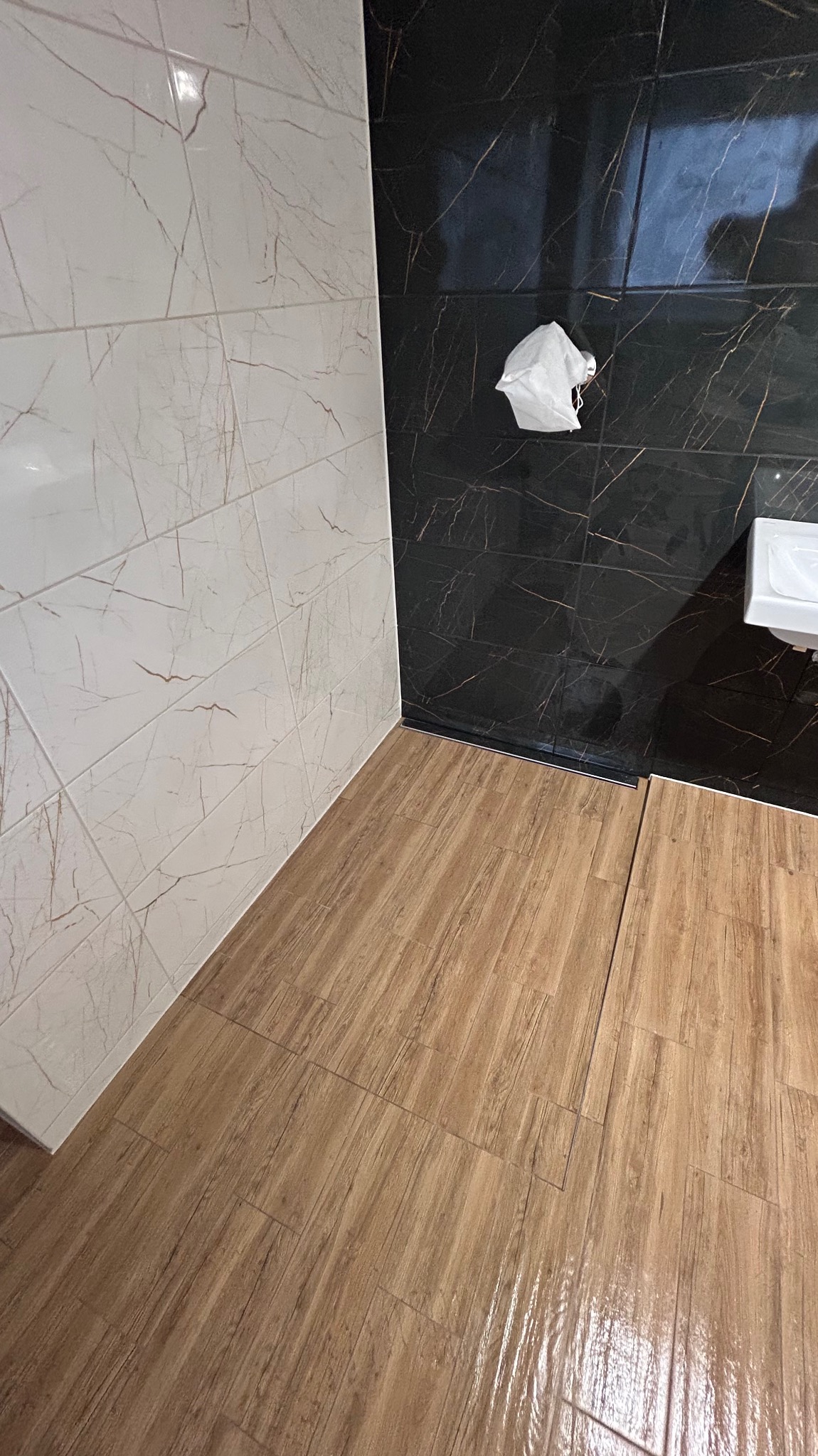 Wykończona łazienka z płytkami imitującymi marmur w odcieniach bieli i czerni, oraz podłogą z paneli imitujących drewno. Widoczny fragment umywalki i akcesoria łazienkowe.