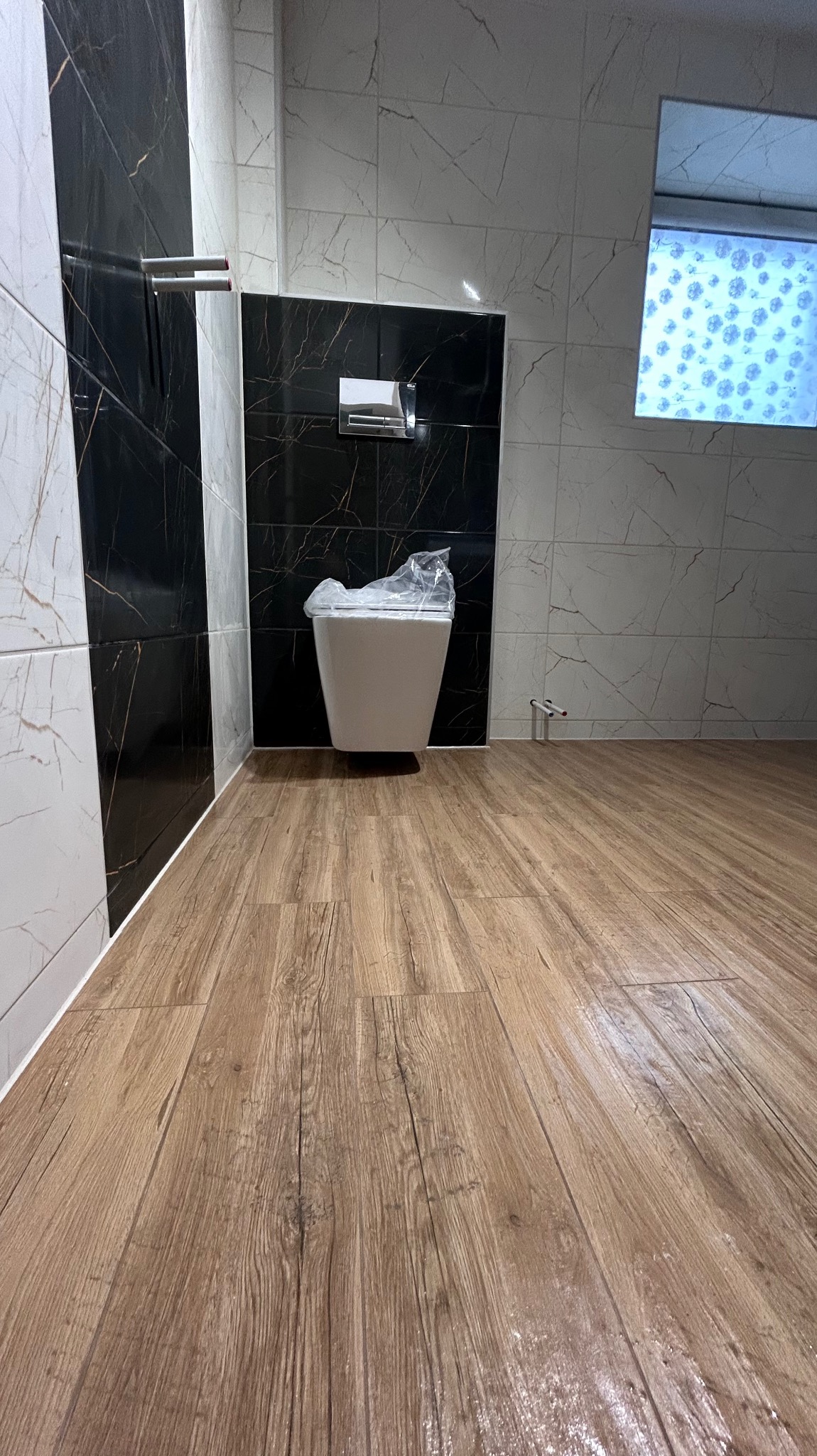 Nowoczesna łazienka z drewnopodobną podłogą, marmurkowymi płytkami i podwieszaną toaletą w folii ochronnej. Minimalistyczny design, świeżo po remoncie.