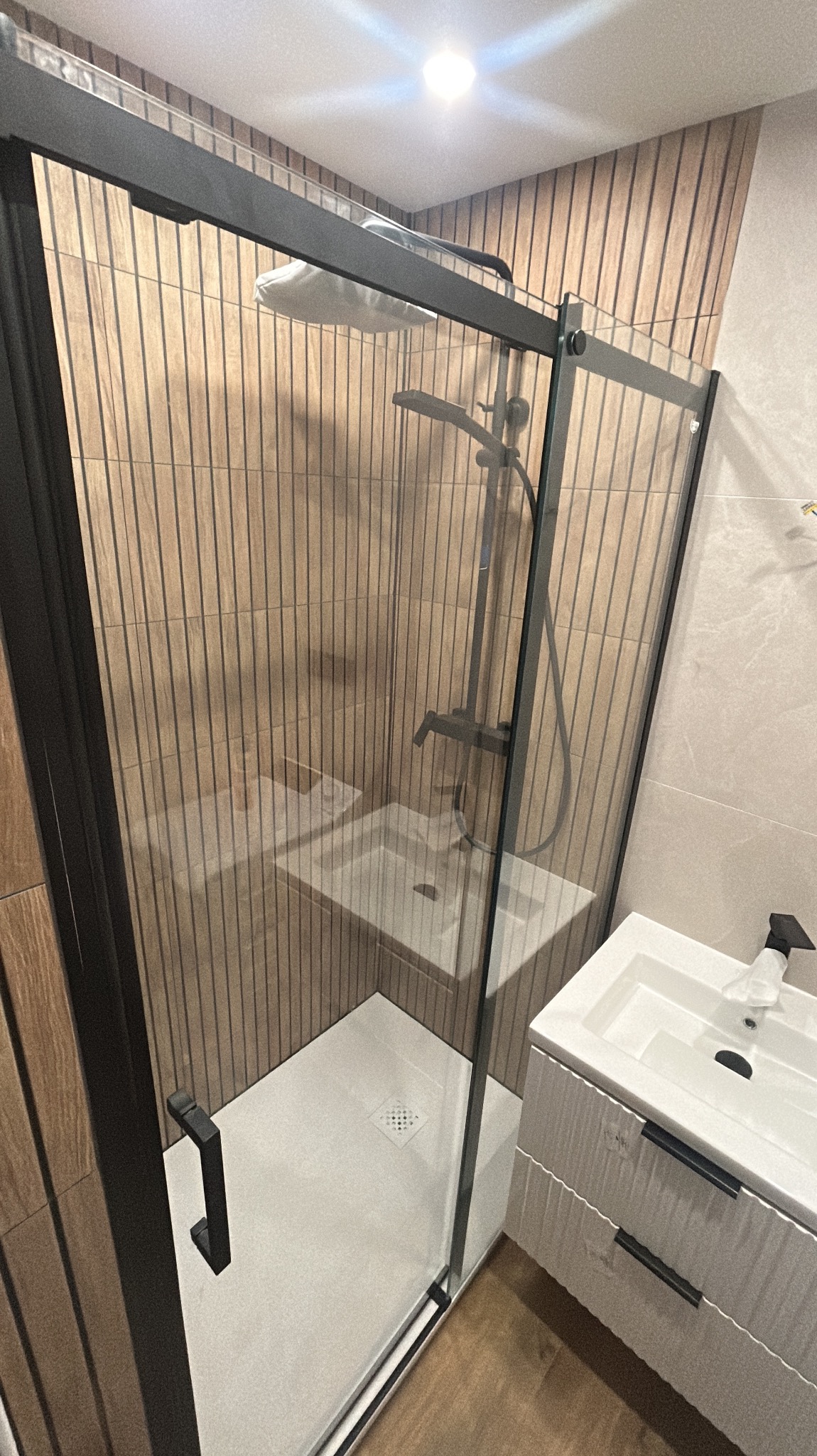 Nowoczesna kabina prysznicowa z czarnymi profilami i deszczownicą, obok umywalka z szafką. Ściany wykończone płytkami imitującymi drewno i jasnymi płytkami.