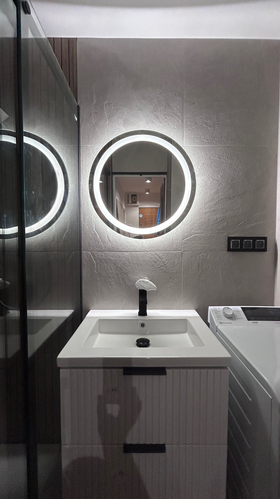 Nowoczesna łazienka z okrągłym lustrem LED, białą umywalką z szafką i pralką. Szare płytki na ścianach. Widoczne elementy kabiny prysznicowej.