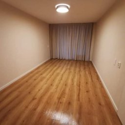 Tommalpol - Pusty pokój po remoncie z panelami podłogowymi w odcieniach brązu, beżowe ściany i zasłony w tle, widoczne listwy przypodłogowe i oświetlenie sufitowe.