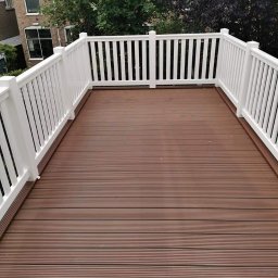 Tommalpol - Nowoczesny taras z brązowych desek kompozytowych i białej balustrady, widok na drzewa i budynek mieszkalny w tle.