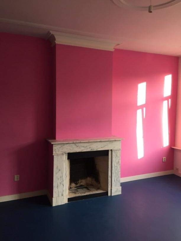 Pokój z pomalowanymi na różowo ścianami, białym marmurowym kominkiem i ciemnoniebieską podłogą, widoczne światło słoneczne padające przez okno.