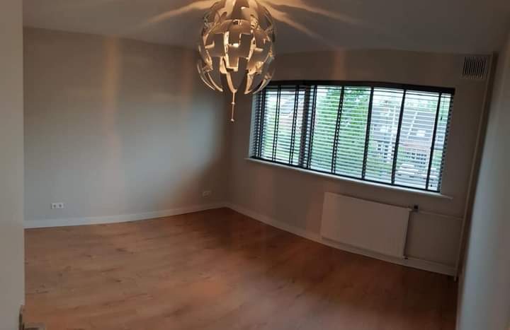 Jasne wnętrze pokoju po remoncie z drewnianą podłogą, szarymi ścianami, nowoczesną lampą i grzejnikiem pod oknem z żaluzjami.