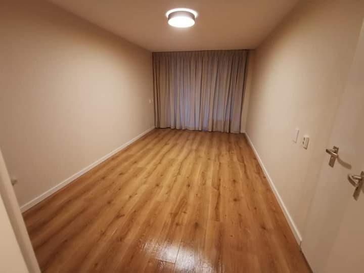 Pusty pokój po remoncie z panelami podłogowymi w odcieniach brązu, beżowe ściany i zasłony w tle, widoczne listwy przypodłogowe i oświetlenie sufitowe.