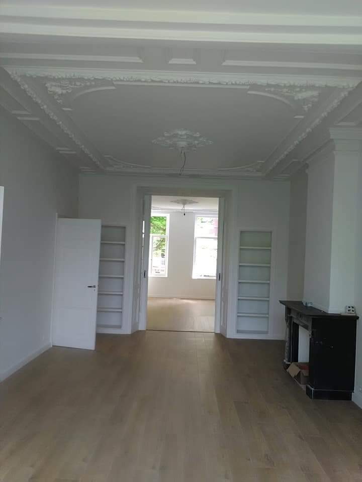 Jasne wnętrze z drewnianą podłogą, ozdobnym sufitem i wnękami na książki, widok na sąsiednie pomieszczenie przez otwarte drzwi.