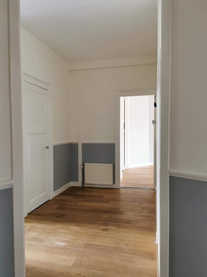 Wnętrze po remoncie: świeżo pomalowane ściany w odcieniach bieli i szarości, ułożona drewniana podłoga, białe drzwi i grzejnik w odnowionym pokoju z widokiem na kolejne pomieszczenie.