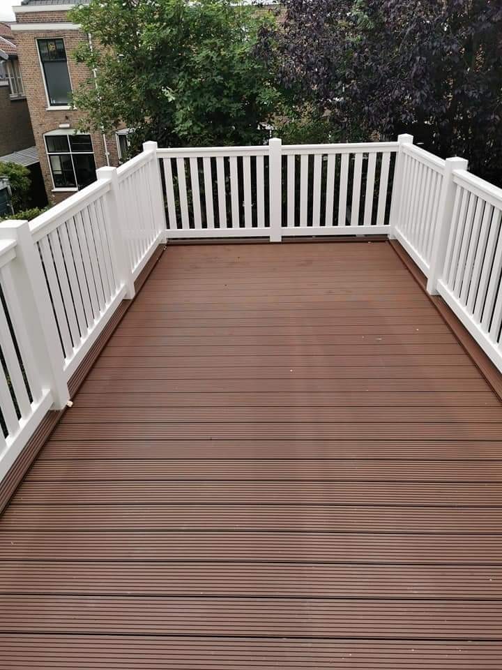 Nowoczesny taras z brązowych desek kompozytowych i białej balustrady, widok na drzewa i budynek mieszkalny w tle.