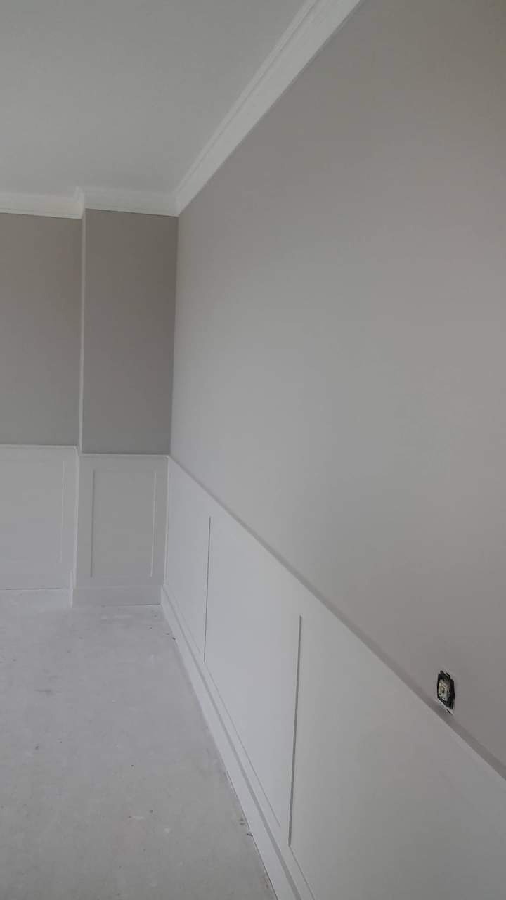 Szara ściana z białą listwą przypodłogową i dekoracyjną boazerią panelową, wykończona białą listwą przysufitową, widoczne gniazdko elektryczne.