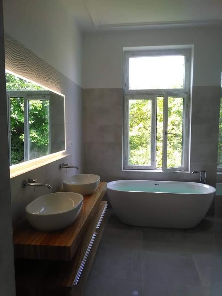Nowoczesna łazienka z owalną wanną wolnostojącą, dwoma umywalkami nablatowymi na drewnianym blacie i dużym oknem z widokiem na zieleń.
