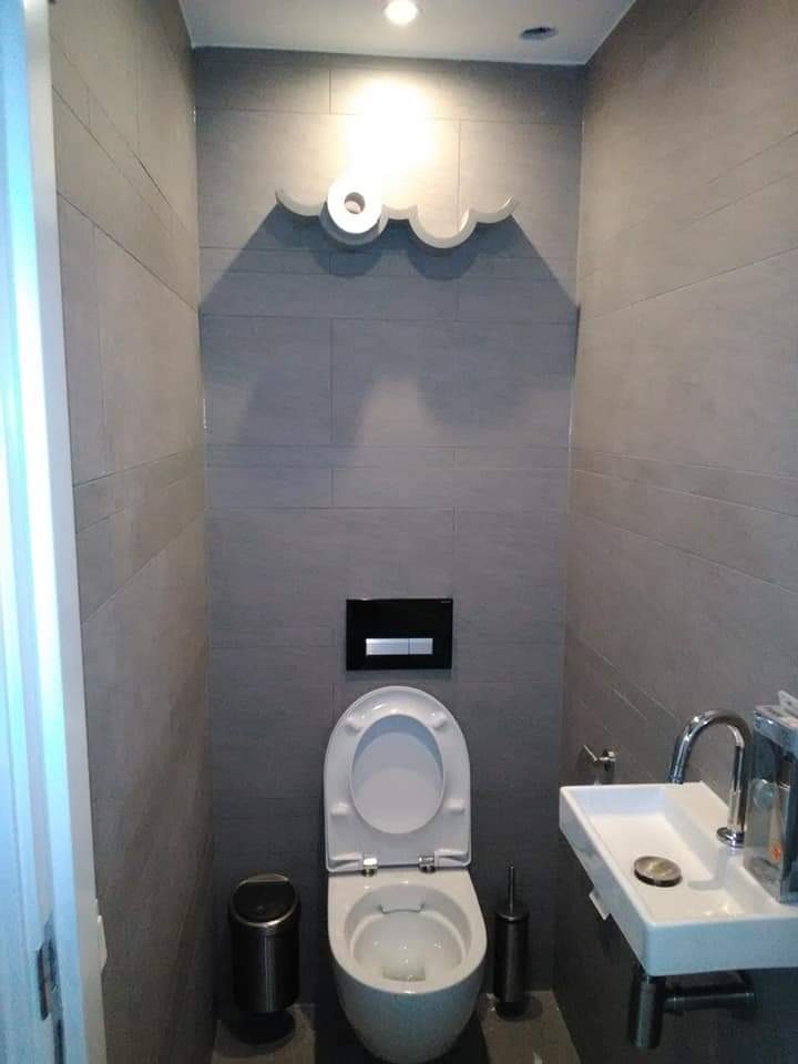 Nowoczesna, wąska łazienka z szarymi płytkami imitującymi cegłę, podwieszana toaleta z otwartą klapą, minimalistyczna umywalka i dekoracyjna półka w kształcie chmury z rolką papieru toaletowego.