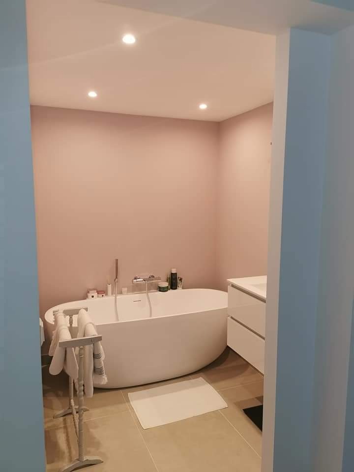 Nowoczesna łazienka z wolnostojącą białą wanną na tle ściany pomalowanej na pudrowy róż, widoczne punktowe oświetlenie sufitowe i biała szafka umywalkowa.