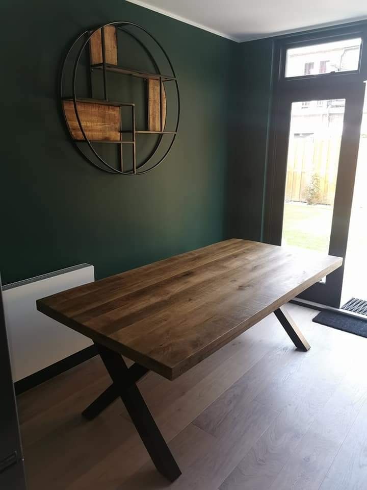 Drewniany stół z czarnymi nogami typu X na jasnej drewnianej podłodze, na ścianie dekoracja w stylu loft z drewnianymi elementami i metalową ramą, ściana pomalowana na ciemnozielony kolor.