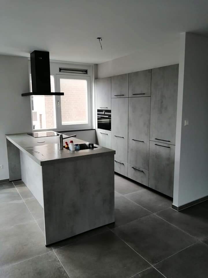 Nowoczesna kuchnia z szarymi szafkami o teksturze betonu, wyspą kuchenną i czarnym okapem, wykończona ciemnymi płytkami podłogowymi.
