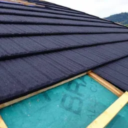 Montaż ciemnoszarej blachodachówki modułowej na drewnianej konstrukcji z widoczną membraną dachową, w tle zalesione wzgórza.