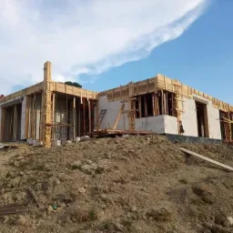 Budowa domu murowanego z betonu komórkowego: ściany w trakcie wznoszenia, szalunki drewniane zamontowane, widok na plac budowy z ziemią i niebem w tle.