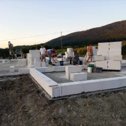 Wznoszenie fundamentów z bloczków betonowych Ytong, widoczni pracownicy budowlani, sprzęt budowlany i malowniczy krajobraz w tle.