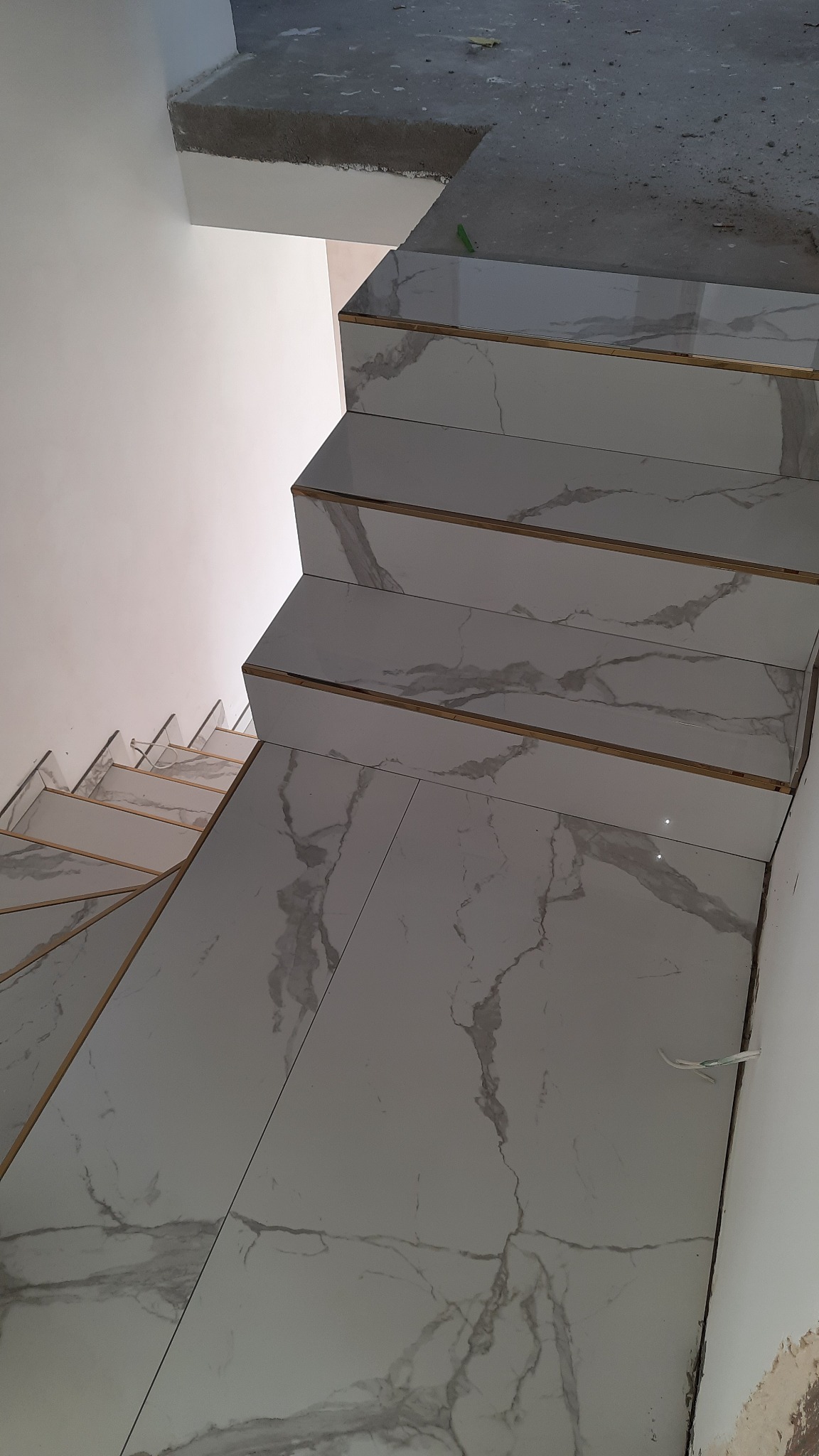 Układanie płytek imitujących marmur na schodach, widoczne złote listwy wykończeniowe, surowy beton na górnym podeście.