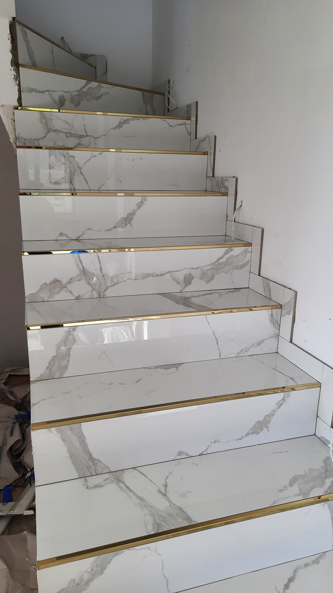 Schody wyłożone białymi płytkami imitującymi marmur z ozdobnymi złotymi listwami, widoczne niedokończone prace wykończeniowe przy ścianie.