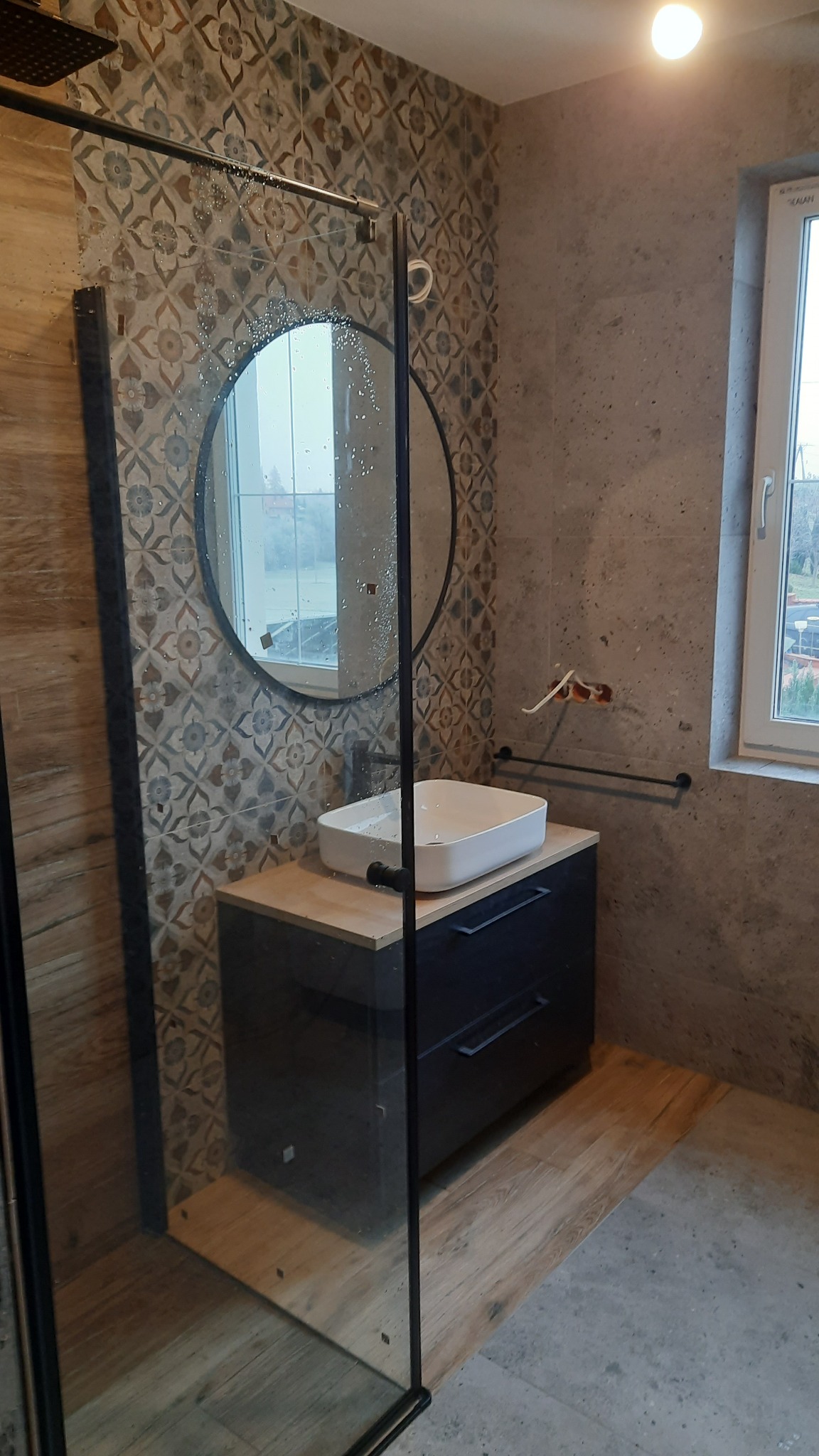 Nowoczesna łazienka z kabiną prysznicową typu walk-in, okrągłym lustrem i umywalką nablatową na czarnej szafce z drewnianym blatem, wykończona płytkami w dwóch wzorach: geometrycznym i imitującym...