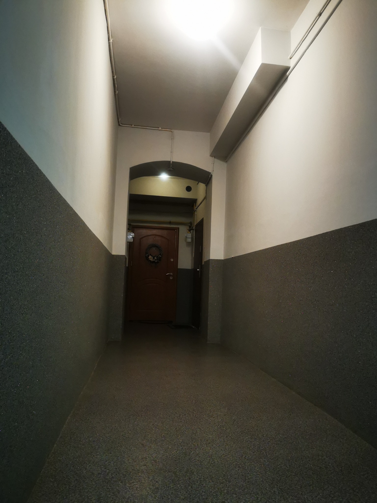 Korytarz w bloku z szarymi ścianami w dolnej części i białymi w górnej, brązowe drzwi z wieńcem w tle, sufit z oświetleniem.