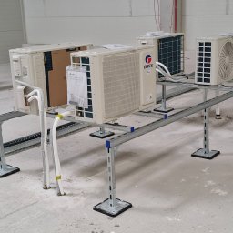 Brat-klim Michał Budz - Instalacja klimatyzacji Gree w hali: cztery jednostki zewnętrzne na metalowej konstrukcji wsporczej, widoczne przewody i elementy montażowe. Hala w surowym stanie.