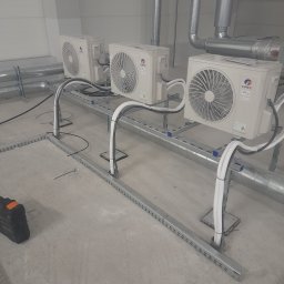Brat-klim Michał Budz - Trzy klimatyzatory marki Gree zamontowane na metalowej konstrukcji w surowym wnętrzu. Widoczne rury i kable, instalacja w trakcie realizacji. Minimalistyczny, techniczny kadr.