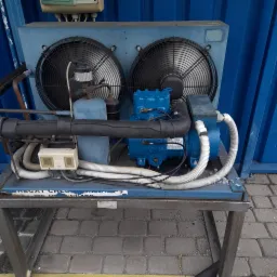 Agregat chłodniczy na metalowym stelażu, z dwoma wentylatorami, rurami i elementami sterującymi, ustawiony na brukowanej powierzchni.