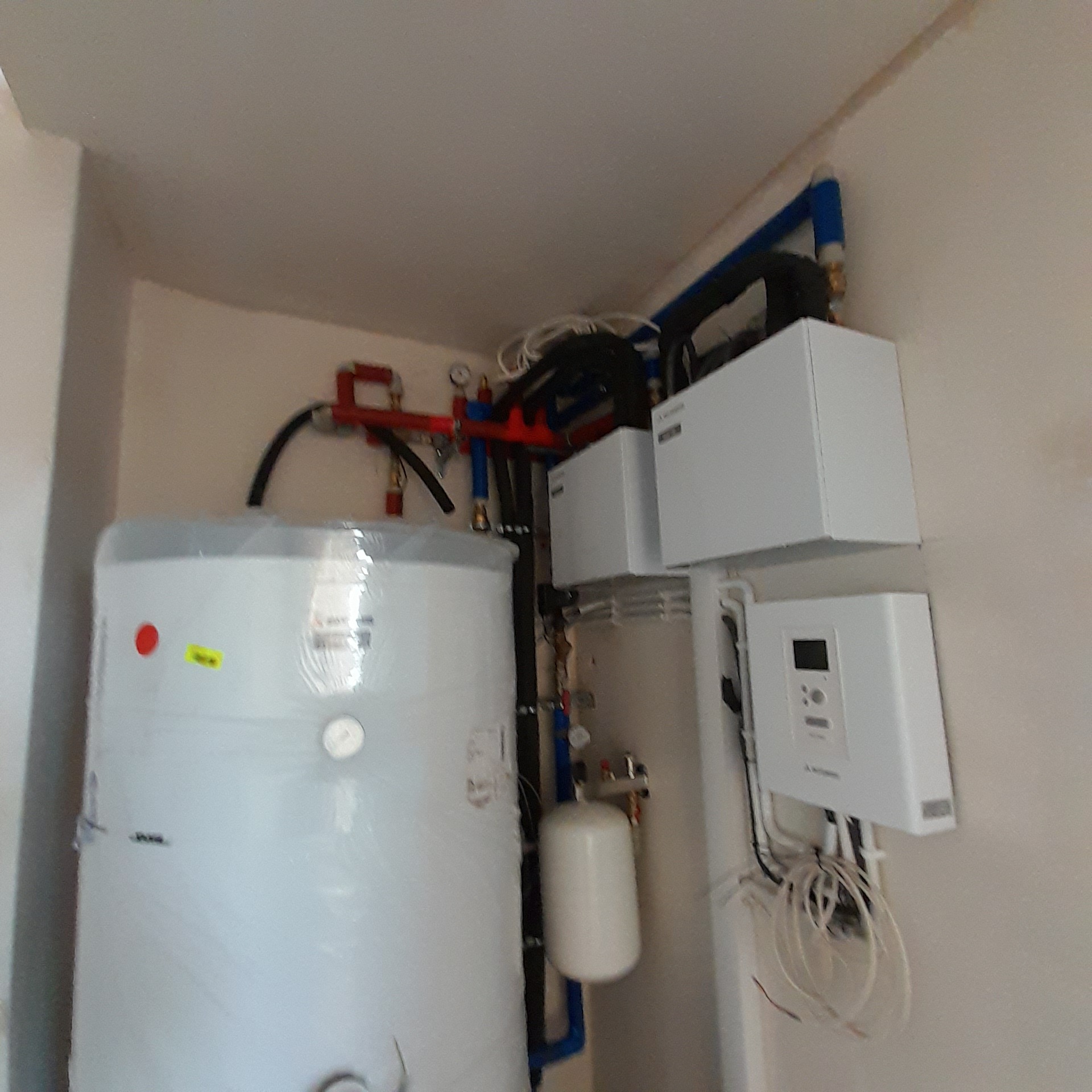 Biały akumulator ciepła owinięty folią, podłączony do instalacji z czerwonymi i niebieskimi rurami, z białymi urządzeniami sterującymi na ścianie, w pomieszczeniu o białych ścianach.