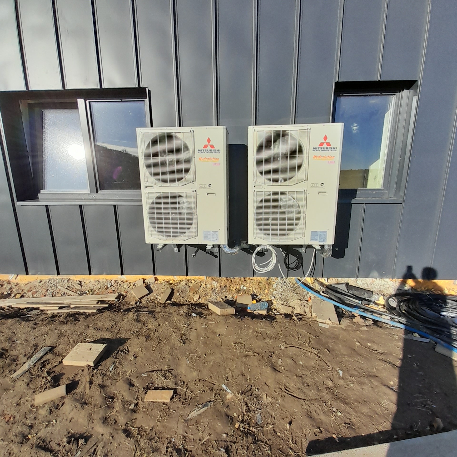 Dwa agregaty klimatyzacyjne Mitsubishi Heavy Industries zamontowane na szarej elewacji budynku z dwoma oknami, widoczne kable i rury, teren wokół w trakcie prac budowlanych.