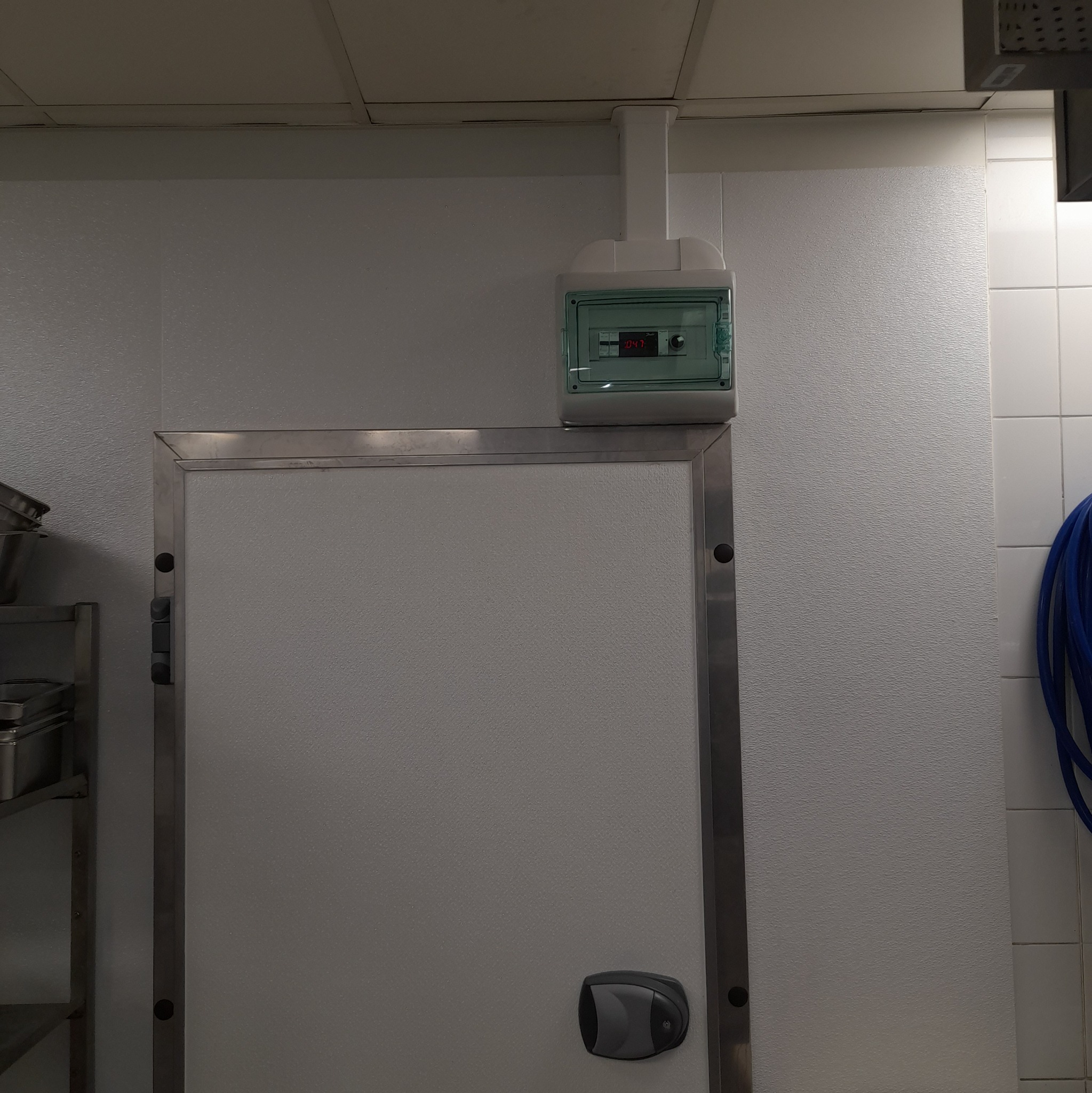 Zamknięte drzwi chłodni z panelem sterowania temperaturą zamontowanym na ścianie. Widoczny fragment węża i metalowego regału.