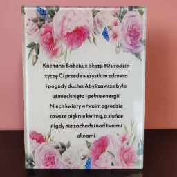 Szklana tablica z życzeniami urodzinowymi dla babci, ozdobiona motywem różowych kwiatów i niebieskich akcentów, ustawiona na ciemnym blacie.