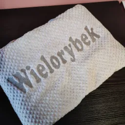 Jasny ręcznik z wytłaczanym wzorem i szarym napisem 'Wielorybek' leżący na ciemnym blacie.