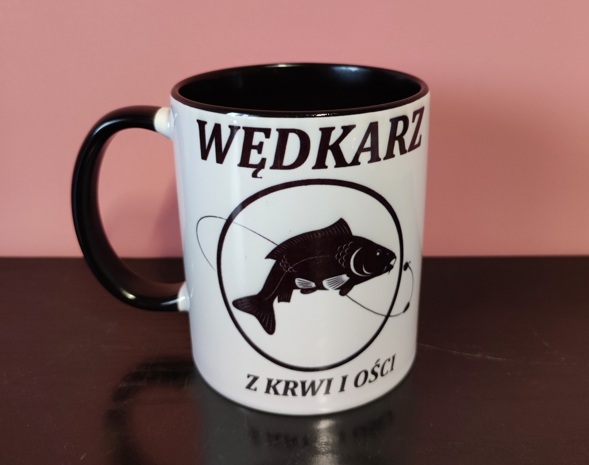 Biały kubek z czarnym wnętrzem i uchwytem, z nadrukiem 'Wędkarz z krwi i ości' oraz wizerunkiem karpia w okręgu z żyłką, na ciemnym blacie i różowym tle.