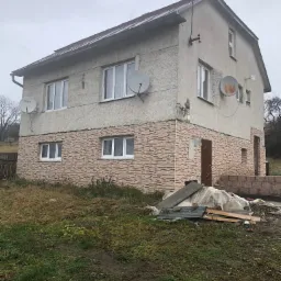 Dwukondygnacyjny dom z ociepleniem dolnej części elewacji imitującym cegłę, z trzema antenami satelitarnymi, widoczne materiały budowlane na zewnątrz.