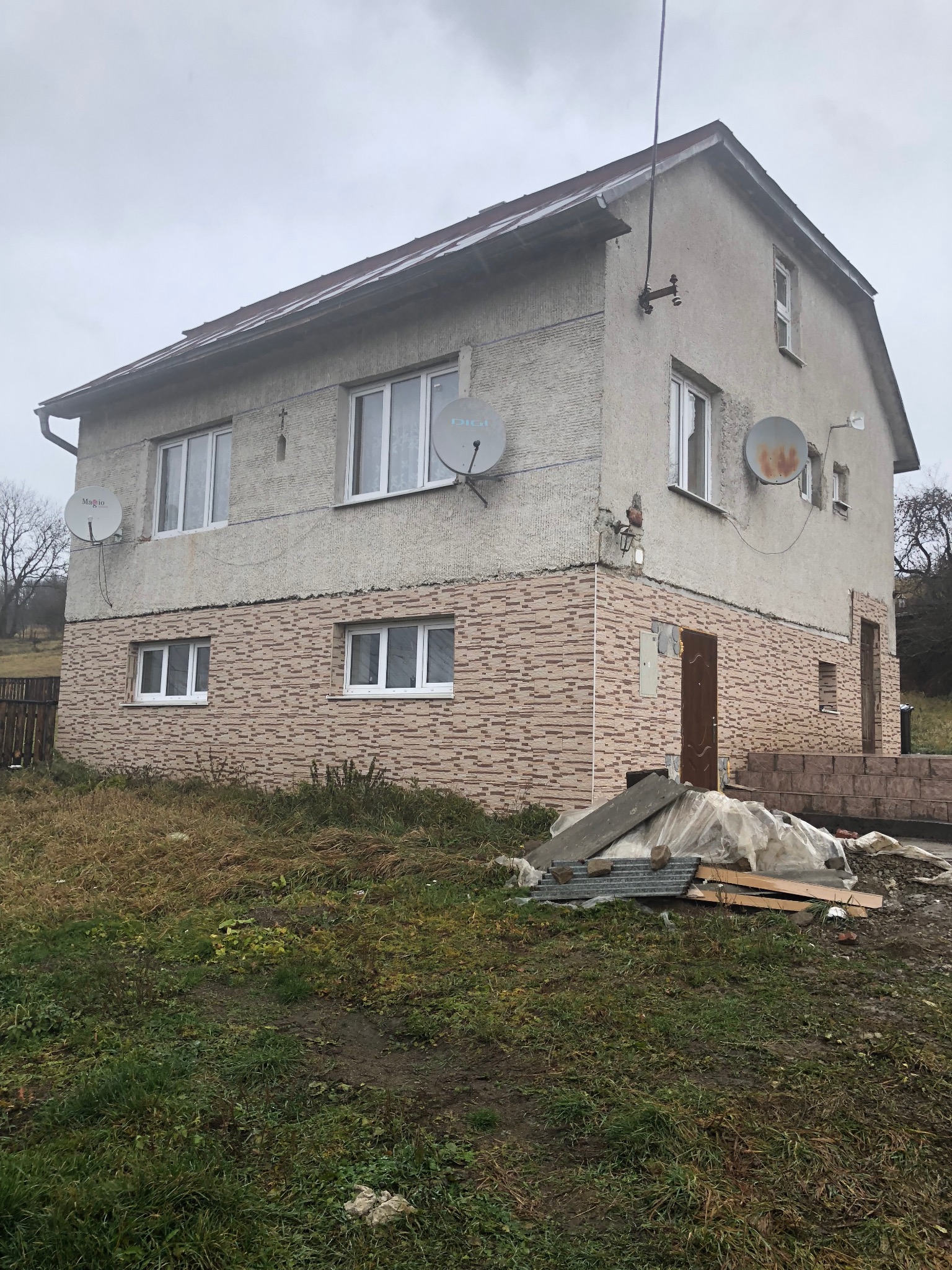 Dwukondygnacyjny dom z ociepleniem dolnej części elewacji imitującym cegłę, z trzema antenami satelitarnymi, widoczne materiały budowlane na zewnątrz.