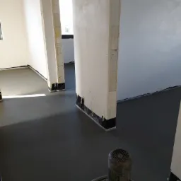 Szlifowanie wylewki w nowym budynku, widoczne przygotowane ściany z taśmą ochronną przy podłodze, szlifierka do posadzek w prawym dolnym rogu.