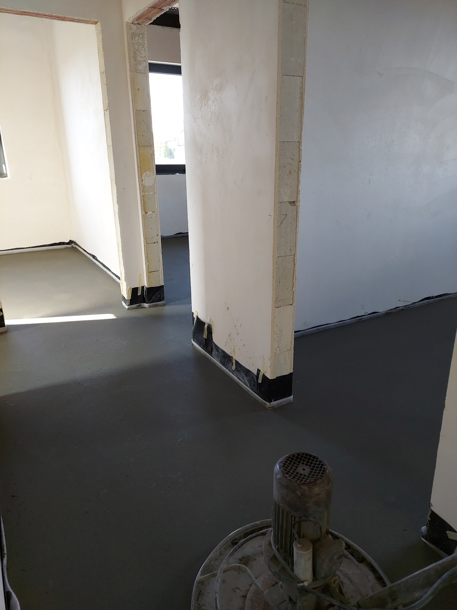 Szlifowanie wylewki w nowym budynku, widoczne przygotowane ściany z taśmą ochronną przy podłodze, szlifierka do posadzek w prawym dolnym rogu.