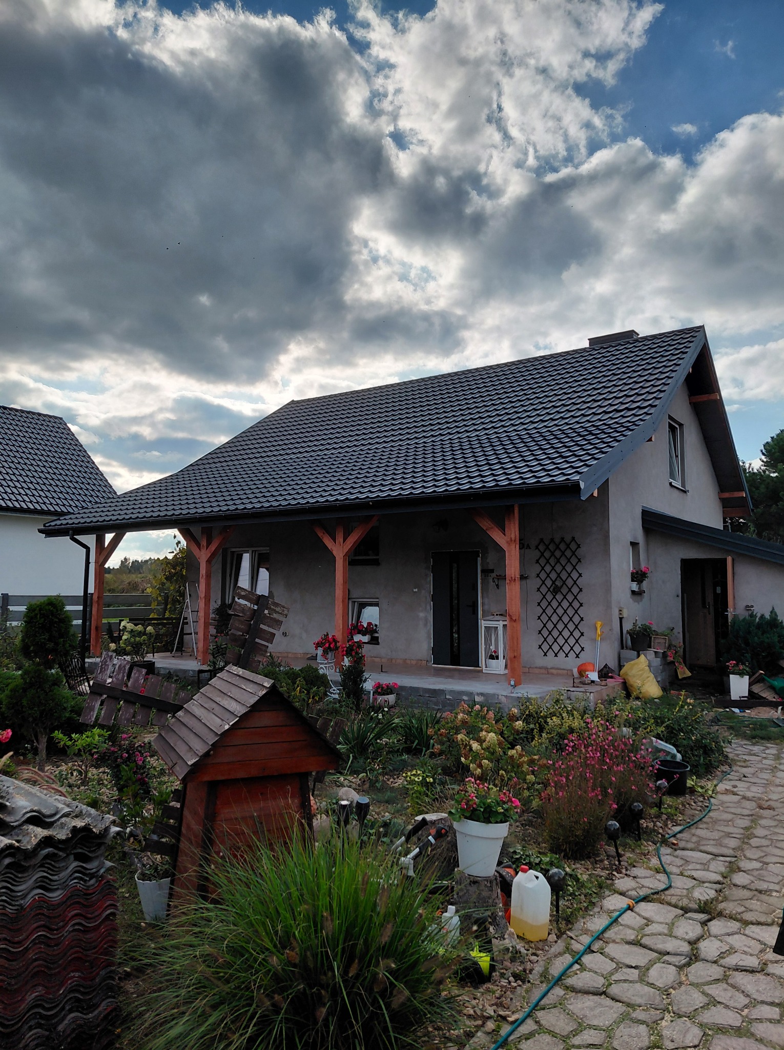 Dom jednorodzinny z ciemną dachówką karpiówką, drewnianymi elementami werandy i bujnym ogrodem. Widoczne stare dachówki, sugerujące wymianę pokrycia.