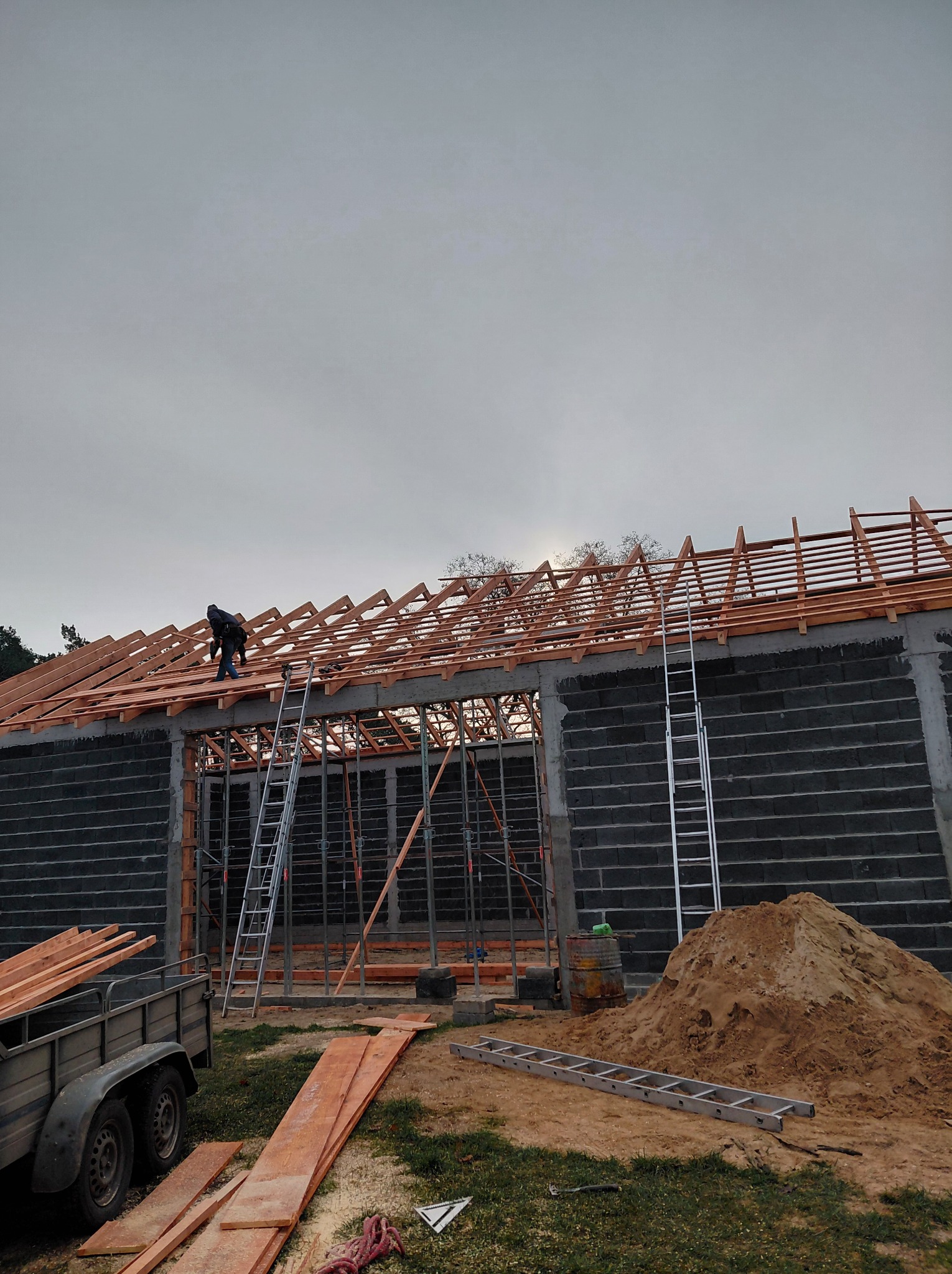 Montaż drewnianej więźby dachowej na budynku z bloczków betonowych. Widoczny pracownik na dachu, drabiny, przyczepka i materiały budowlane. Pochmurne niebo w tle.